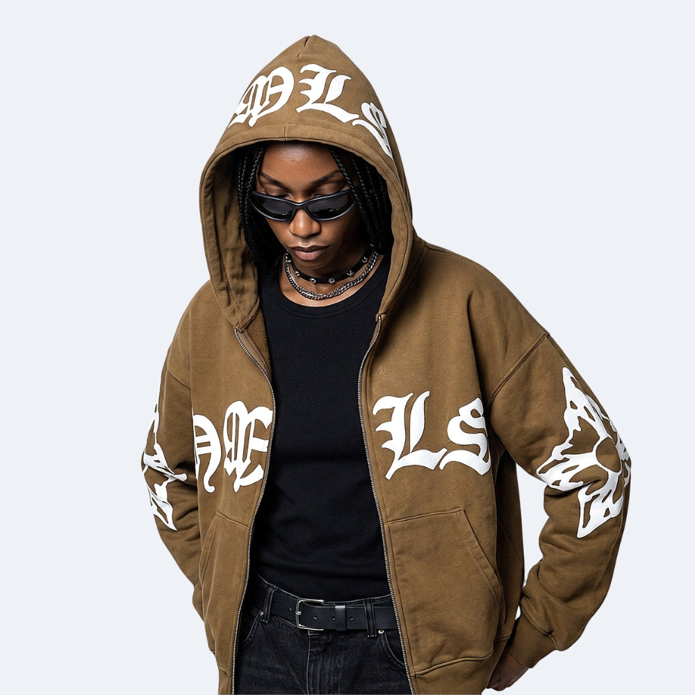 Brown. A person wearing an oversized brown zip-up hoodie with large white Gothic-style lettering on the hood, sleeves, and front, styled over a black top and jeans.