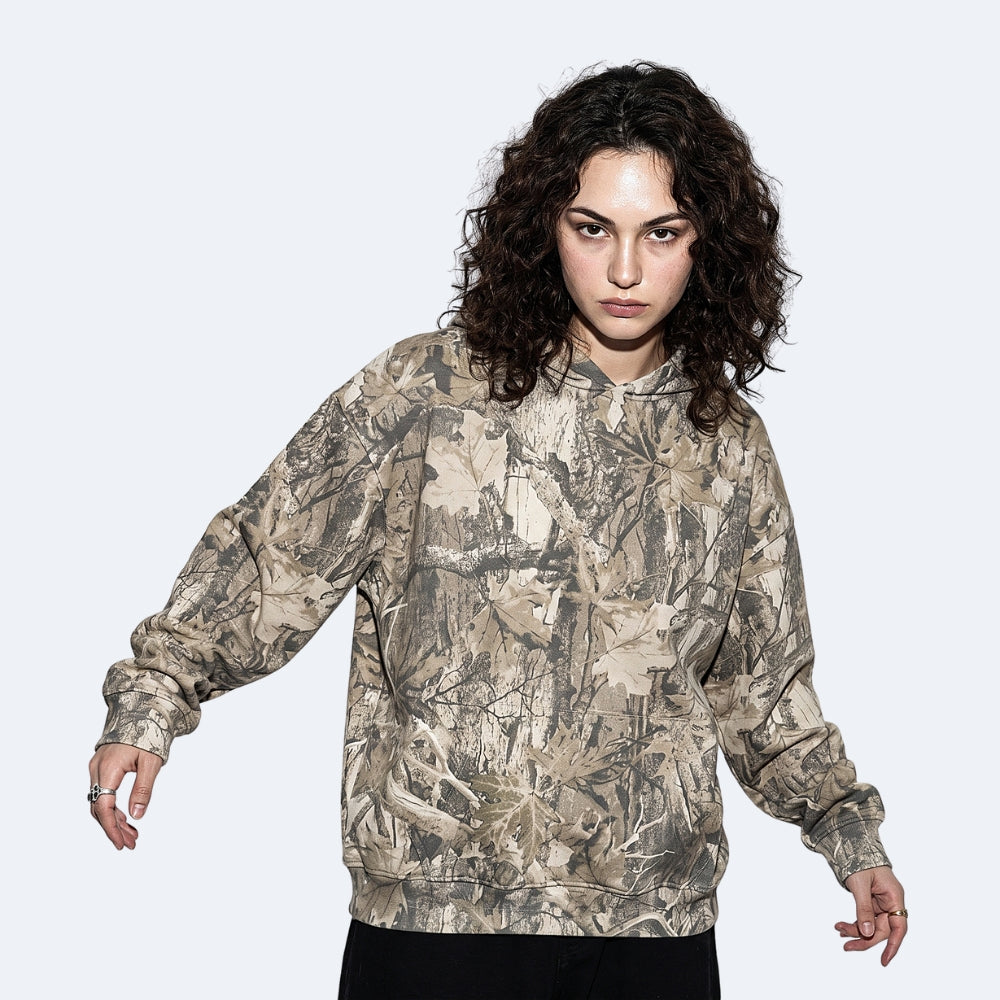 Birch. A model with curly dark hair wearing a camouflage hoodie with a realistic leaf and branch pattern in shades of beige, brown, and gray.