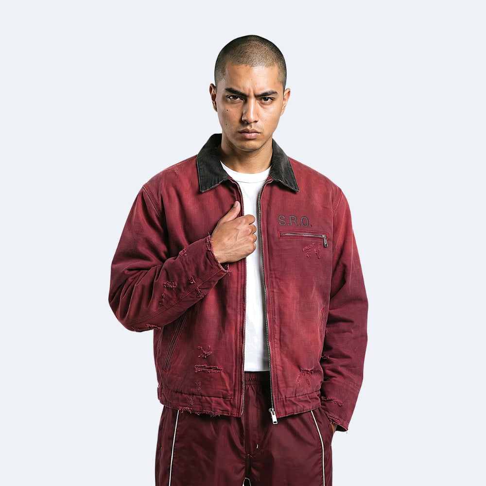 A man wearing a distressed maroon bomber jacket with a black corduroy collar, a white t-shirt, and matching maroon pants, standing against a light gray background.