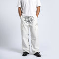 A person wearing off-white cargo pants with a large gray graphic print on the left thigh and a small logo on the right pocket, paired with a white t-shirt and black loafers, shown from the chest down against a light gray studio background.