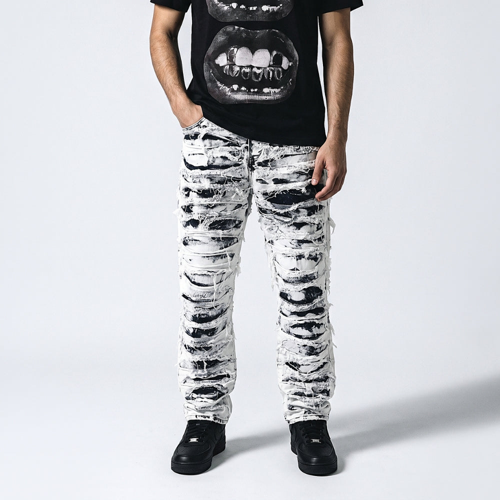 A person wearing distressed black and white ripped jeans with frayed edges, paired with a black graphic t-shirt and black shoes, standing against a light gray studio background.
