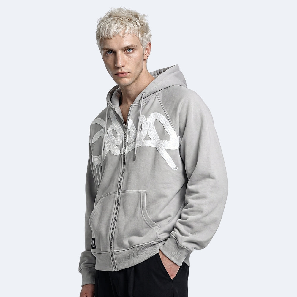 A man with short blonde hair wearing a light gray zip-up hoodie with a large white graphic print on the front, paired with black pants, set against a solid light gray background.