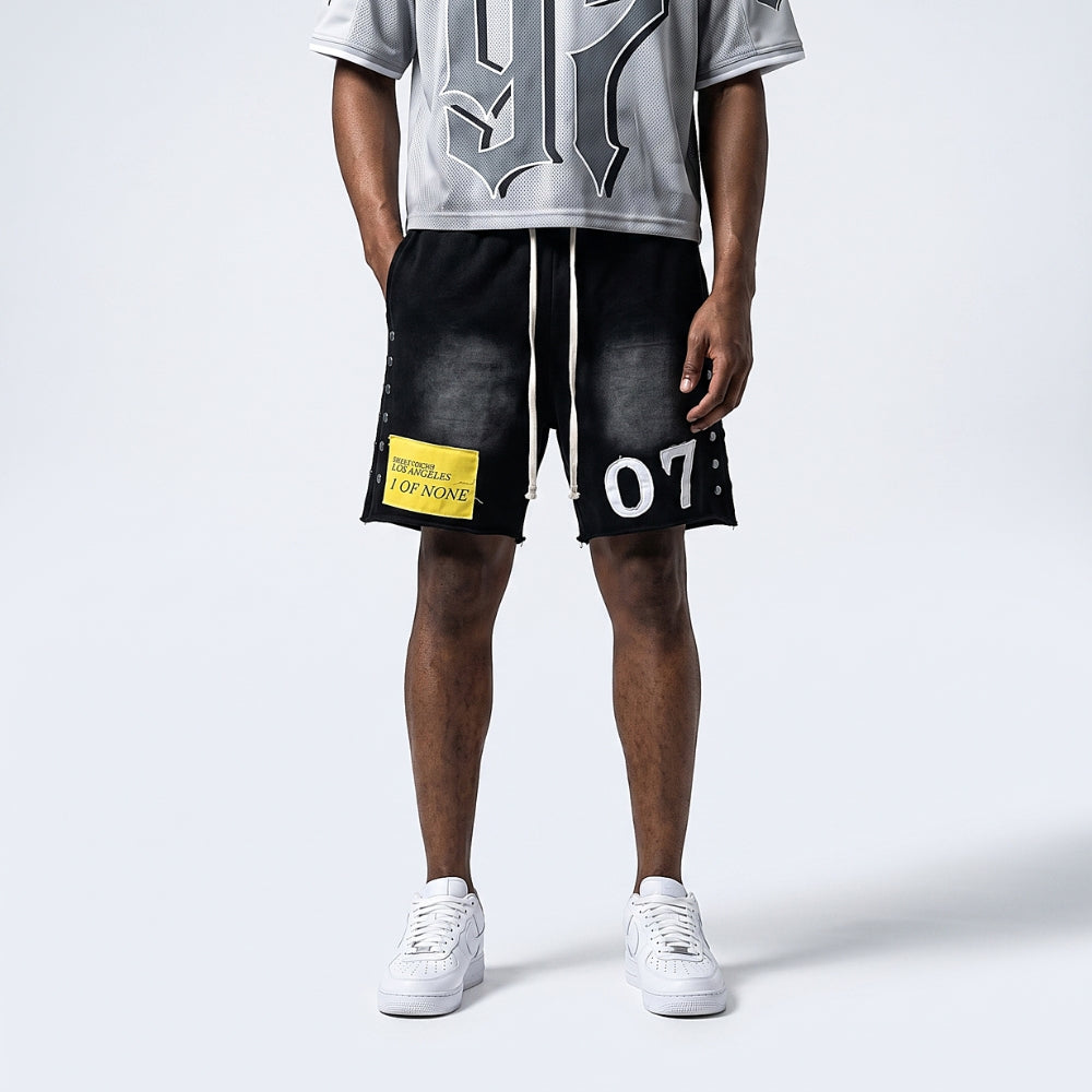 A model is shown from the waist down wearing black washed athletic shorts with white drawstrings, a yellow 'TERRITORY OF THE LAP NINE' patch on the left leg, and white sneakers, paired with a gray graphic t-shirt against a light gray studio background.