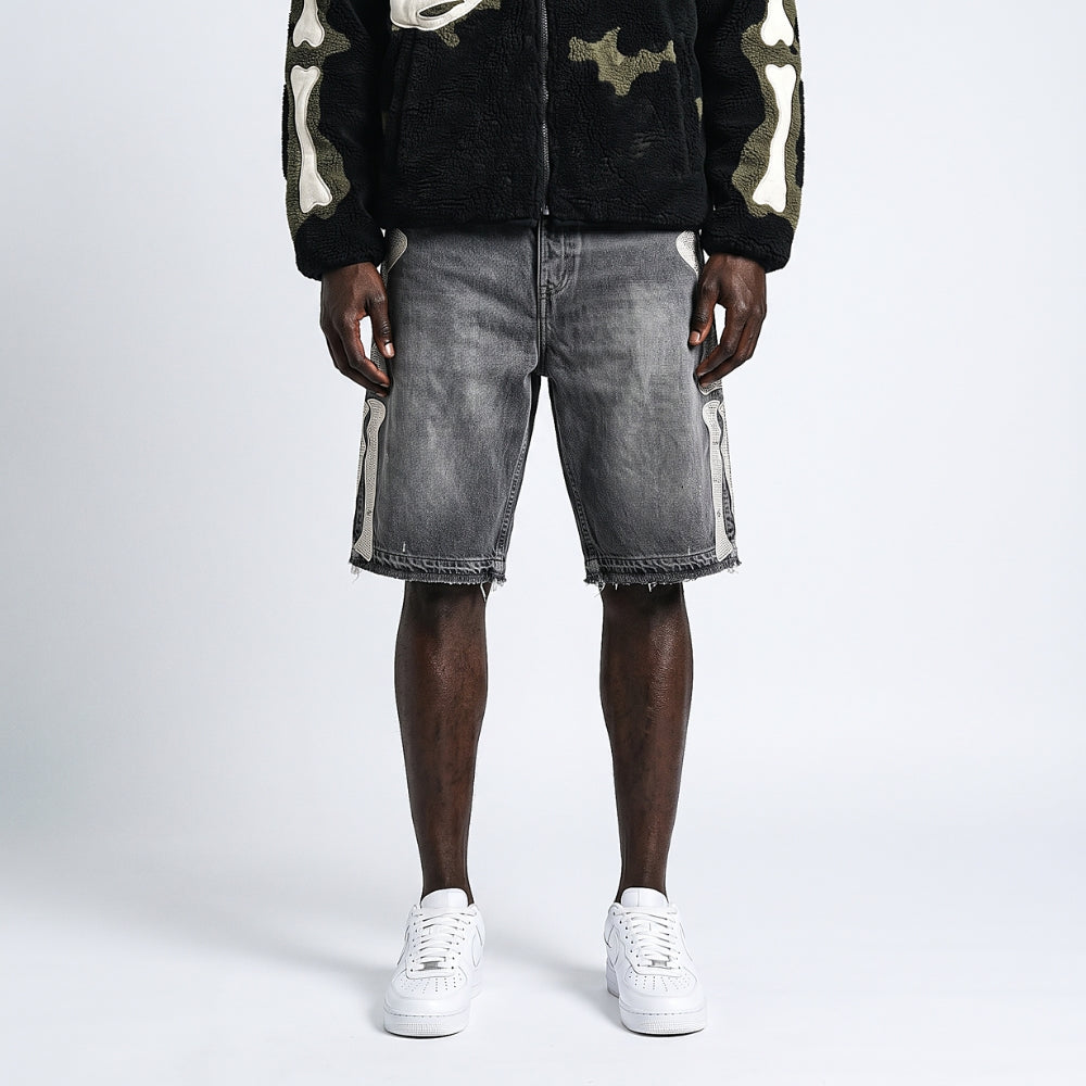 A model is shown from the chest down wearing faded gray denim shorts with white side stripes, a black fleece zip-up jacket with camouflage sleeves, and white sneakers, set against a plain light gray studio background.