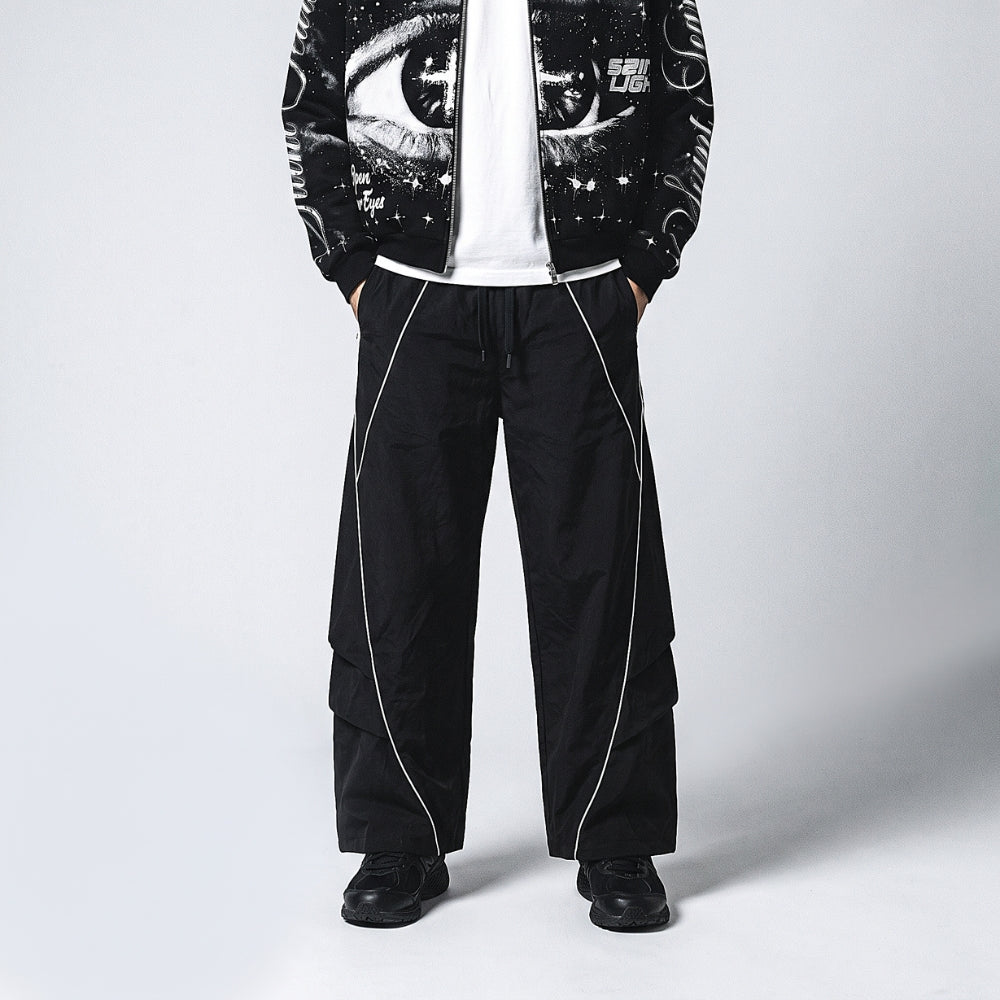 A model wearing black wide-leg cargo pants with white piping, a white t-shirt, and a black graphic zip-up jacket against a light gray studio background.