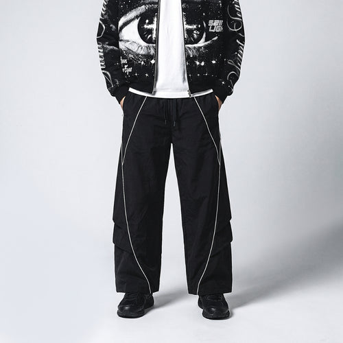 A model wearing black wide-leg cargo pants with white piping, a white t-shirt, and a black graphic zip-up jacket against a light gray studio background.
