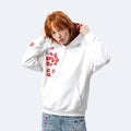 A model with red hair wearing a white hoodie decorated with red lipstick kiss prints, paired with light blue jeans featuring red paint splatters, set against a solid light gray background.