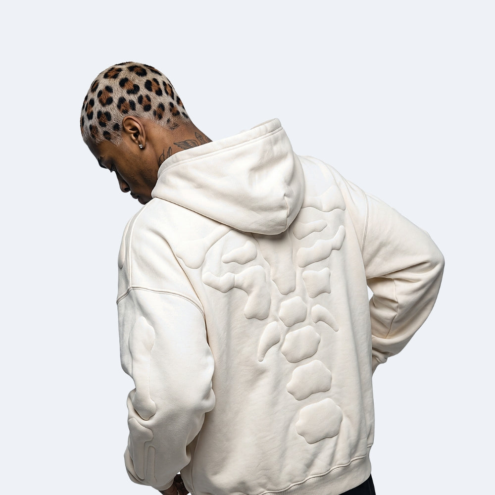 White. A man wearing a cream-colored hoodie with a large 3D abstract bone design on the back, shown from behind.