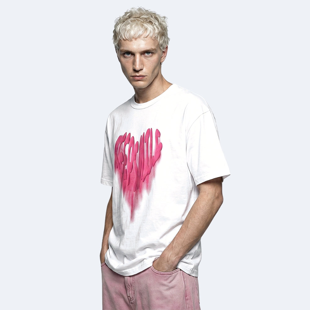 A man with platinum blonde hair wearing a white t-shirt with a pink dripping heart graphic and pink pants, standing against a light gray background.