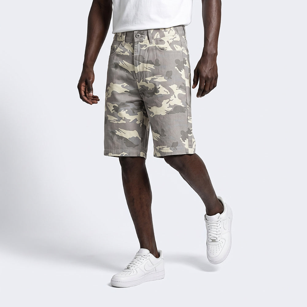 A model wearing beige and gray camouflage cargo shorts, styled with a white t-shirt and white sneakers, set against a plain light gray studio background.