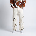A person wearing cream-colored denim jeans with black and brown abstract graphic prints, paired with a brown and white graphic zip-up jacket and white sneakers, shown from the chest down on a light gray studio background.