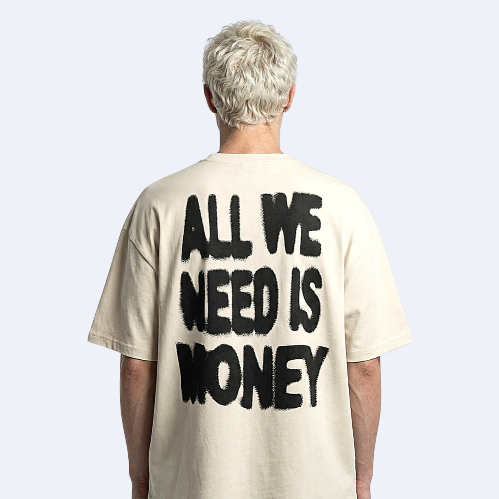 A person viewed from the back wearing an oversized cream-colored t-shirt with the phrase 'ALL WE NEED IS MONEY' printed in a large, black, distressed brush-stroke font on the back.