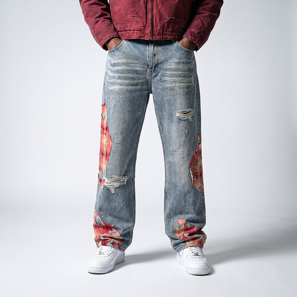 A person wearing light-wash distressed denim jeans with red and orange floral patches on the thighs and lower legs, paired with a burgundy jacket and white sneakers, photographed against a light gray studio background.