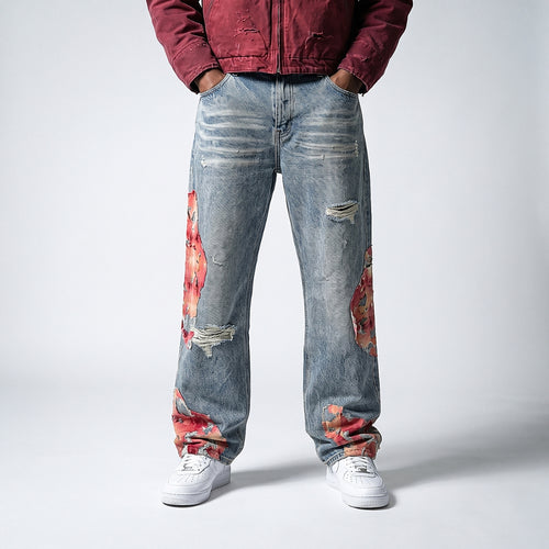 A person wearing light-wash distressed denim jeans with red and orange floral patches on the thighs and lower legs, paired with a burgundy jacket and white sneakers, photographed against a light gray studio background.