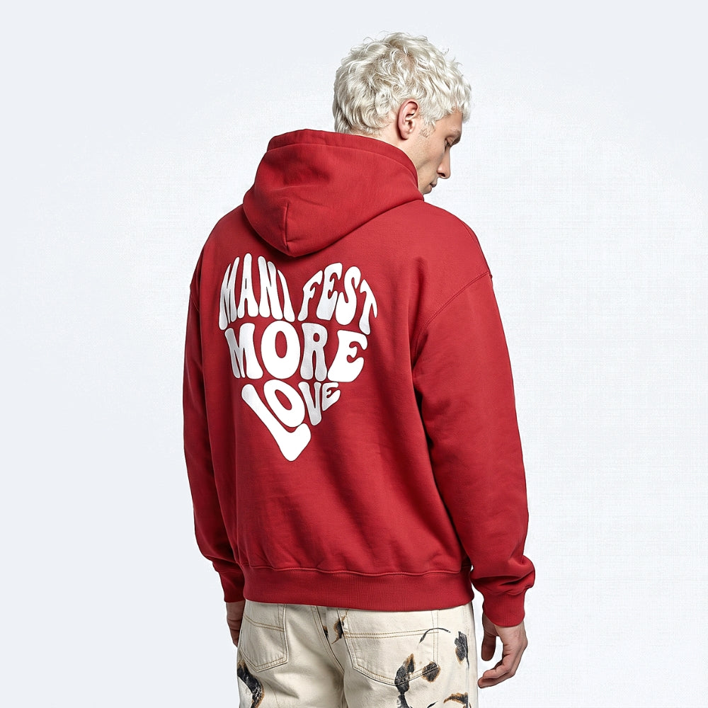 Red. A man with platinum blonde hair shown from behind wearing a red hoodie with a large white graphic on the back that reads MANIFEST MORE LOVE.