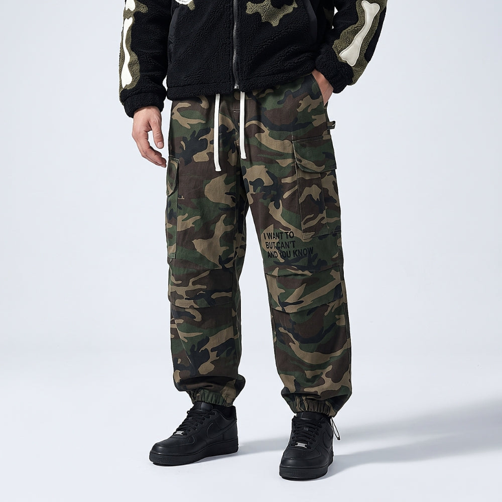 A person wearing camouflage jogger pants with a black fleece jacket and black sneakers, standing against a light gray studio background.