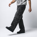 A person wearing black denim jeans with large, abstract white embroidered or printed patterns, paired with a light gray graphic t-shirt and black sneakers, captured in a side-profile walking pose against a light gray studio background.