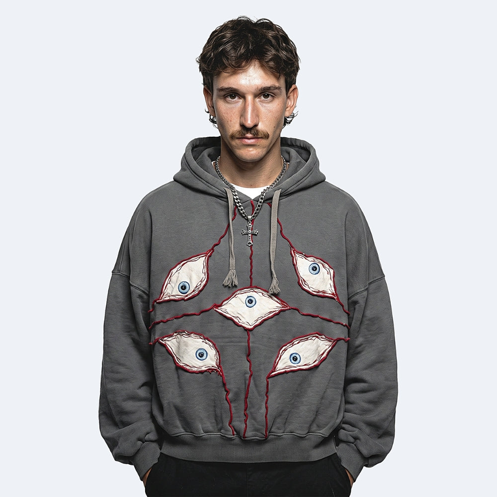 A man wearing a dark gray pullover hoodie with a graphic design of four stylized eyes connected by red lines, photographed against a light gray studio background.