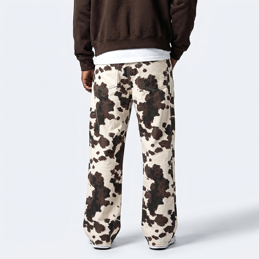 A person shown from the back wearing loose-fit pants with a brown and cream cow-print pattern, a dark brown sweatshirt, and a white undershirt, against a light gray studio background.