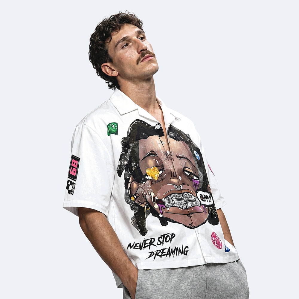 A man wearing a white short-sleeve button-up shirt with a large multicolor graphic print of a face and the text 'NEVER STOP DREAMING' on the front, paired with gray sweatpants against a light gray background.