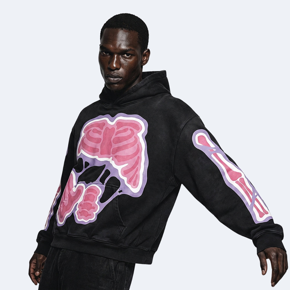 A man wearing a black pullover hoodie with a pink and purple anatomical rib cage graphic on the chest and a matching skeletal arm graphic, posed against a light gray background.
