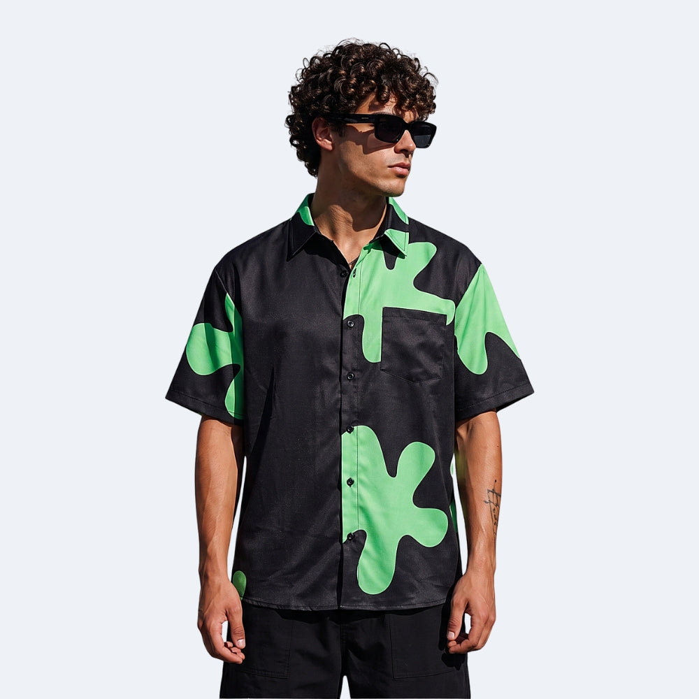 Black & Green. A man wearing a black short-sleeve button-up shirt with a bright green abstract camouflage pattern, styled with black sunglasses.