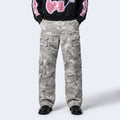 A person wearing loose-fit camouflage cargo pants with multiple flap pockets, paired with a black hoodie featuring pink and white graphic details on the sleeves and upper chest, and black shoes, photographed against a light gray studio background.