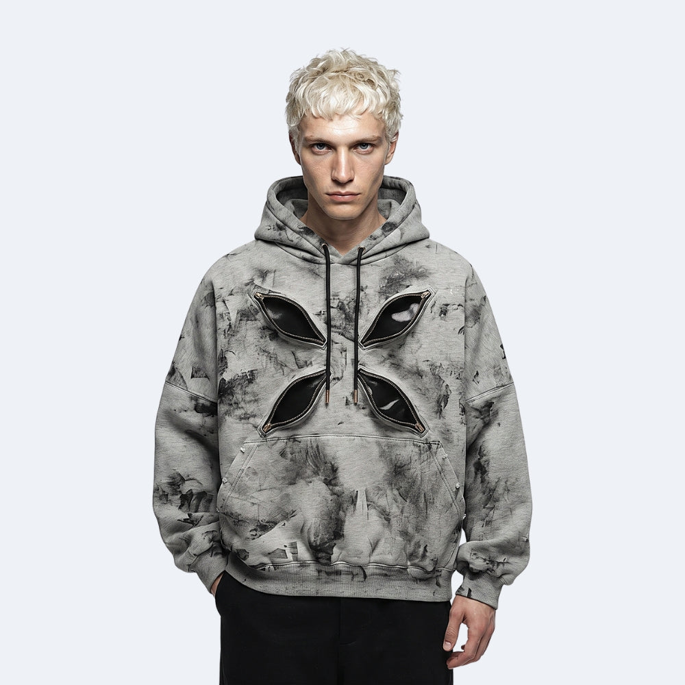 A model with platinum blonde hair wearing a gray and black marbled hoodie with a large black geometric cross graphic on the chest, set against a plain light gray background.
