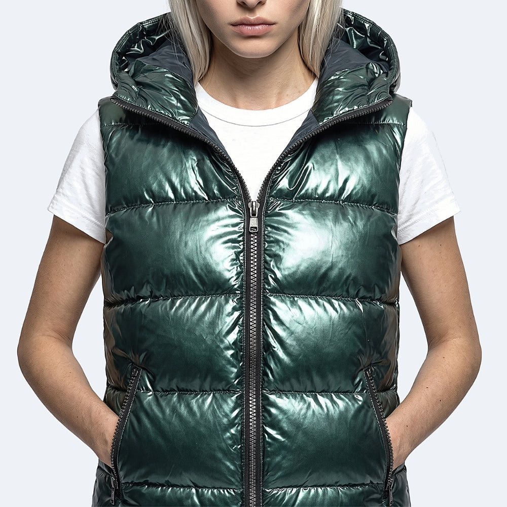 A woman wearing a shiny dark green puffer vest over a white t-shirt, shown from the neck down against a light gray background.