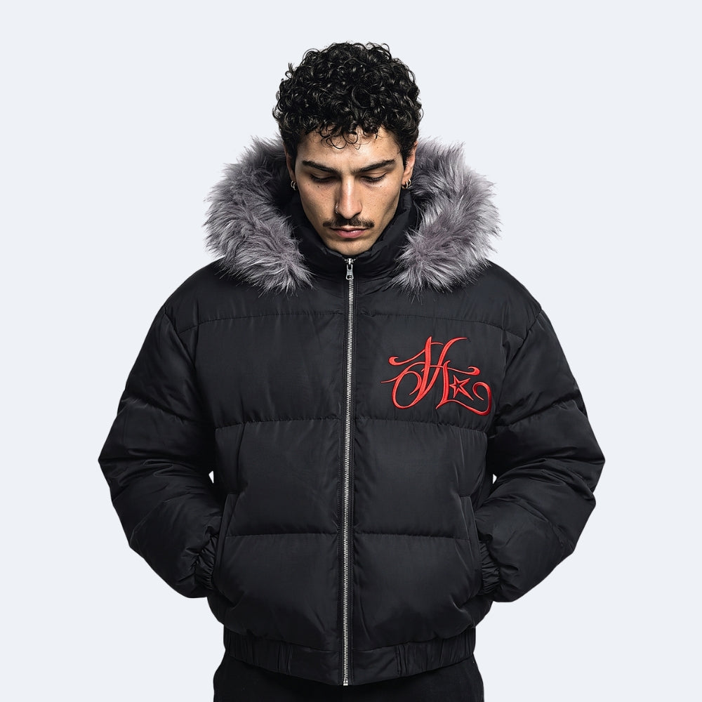 A man wearing a black puffer jacket with a gray faux-fur trimmed hood and a red embroidered logo on the chest, set against a light gray background.