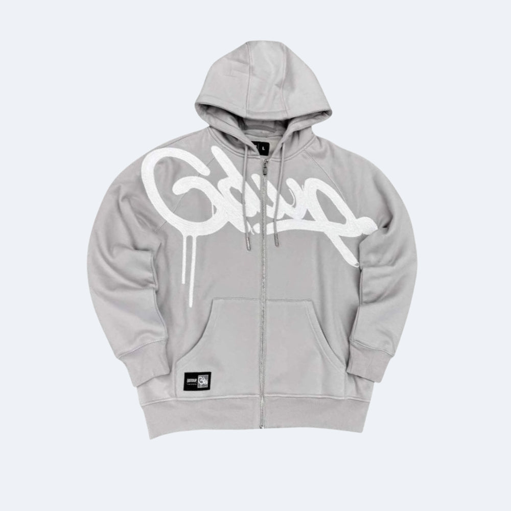 A light gray zip-up hoodie with a large white graffiti-style graphic across the chest, featuring a front kangaroo pocket and drawstrings, shown against a light gray background.