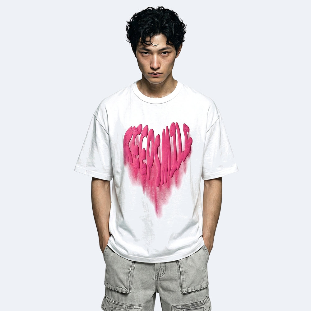 A man wearing a white crewneck t-shirt with a large pink graffiti-style heart graphic on the front, paired with light gray cargo pants, against a light gray background.