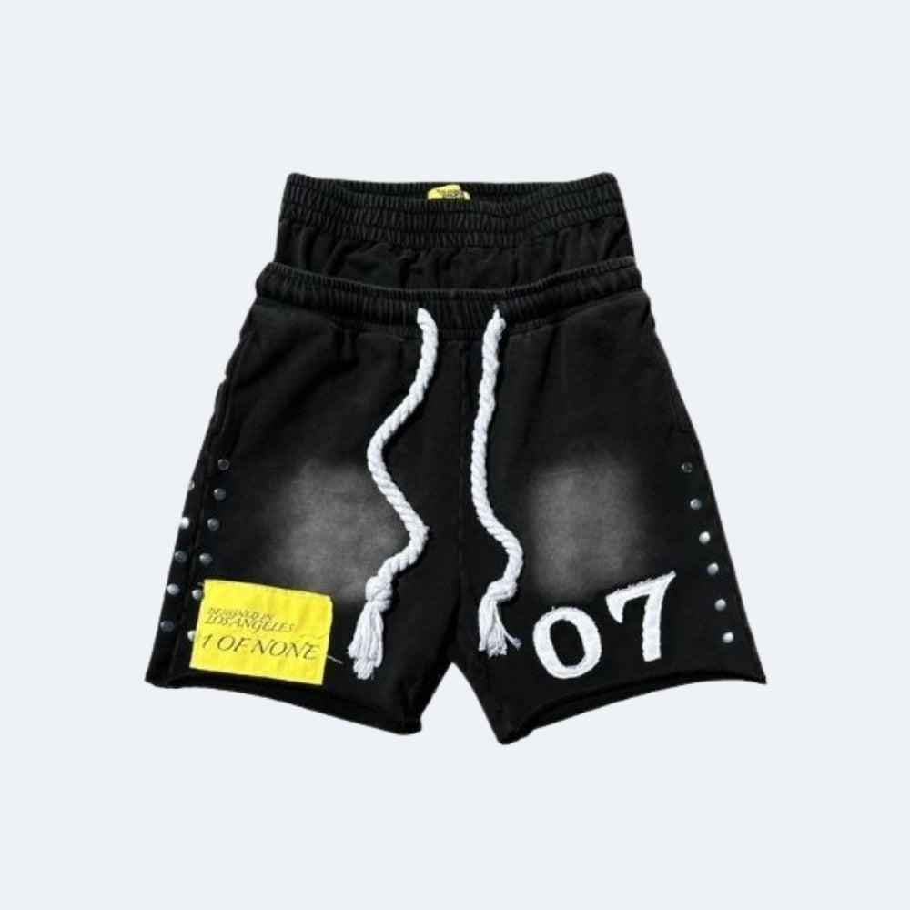 Black washed athletic shorts with white rope drawstrings, white '07' text on the left leg, silver rivet details along the sides, and a yellow patch on the right leg, shown against a light gray background.