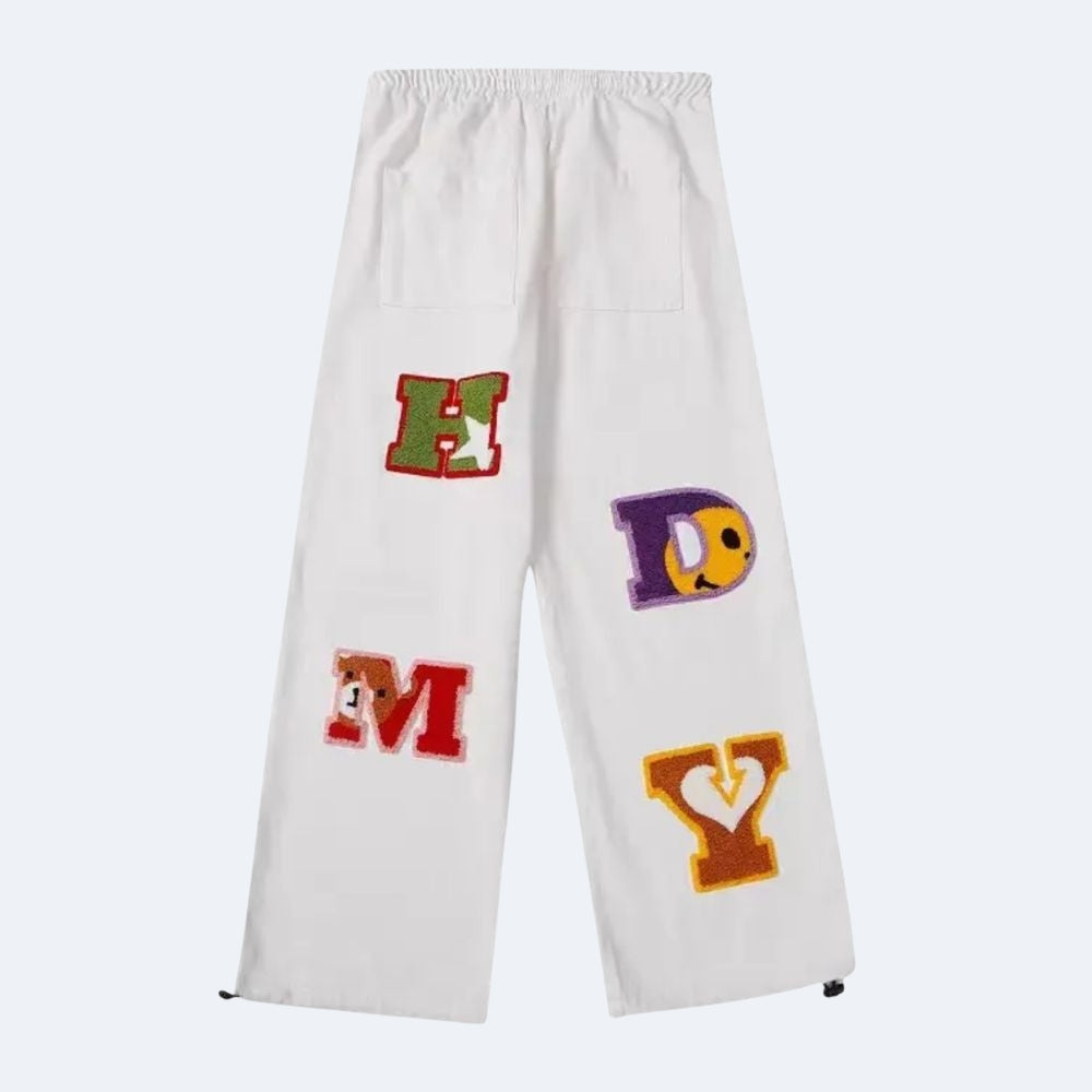 White. White cropped sweatpants with an elastic waistband and pockets, featuring colorful embroidered varsity-style letters on the legs.
