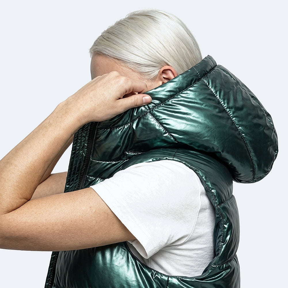 A person with platinum blonde hair wearing a shiny metallic dark green puffer vest over a white t-shirt, shown from the side and back against a light gray background.