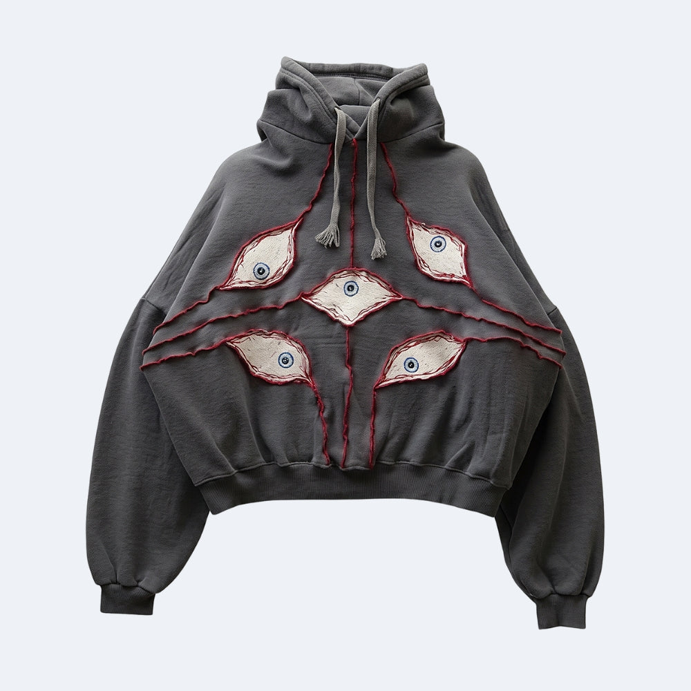 A dark gray cropped hoodie featuring five stylized embroidered eyes with red stitched outlines and blue centers across the front, displayed on a light gray background.