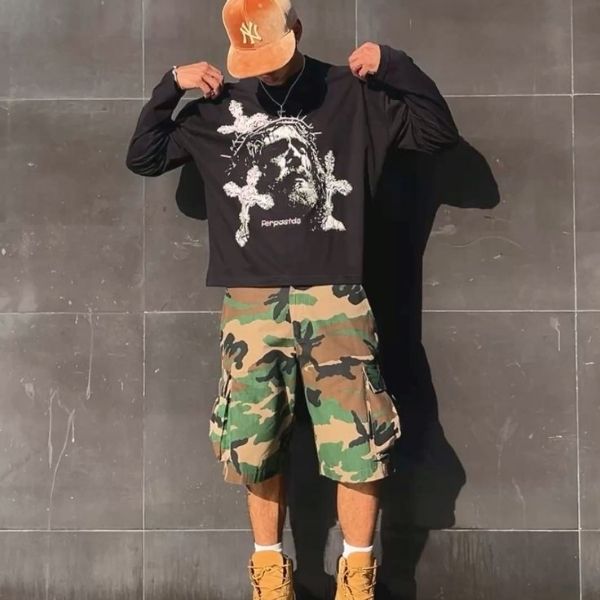 Person wearing a black graphic sweatshirt, camouflage shorts, and tan boots against a dark tiled wall.