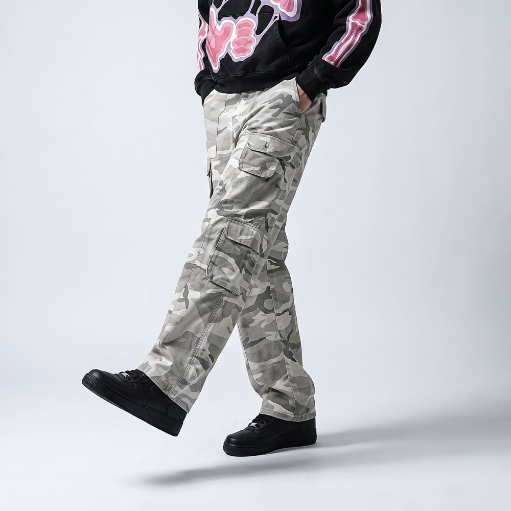 A person wearing light gray and white camouflage cargo pants, styled with a black graphic sweatshirt and black sneakers, captured in a side-profile walking pose against a light gray studio background.