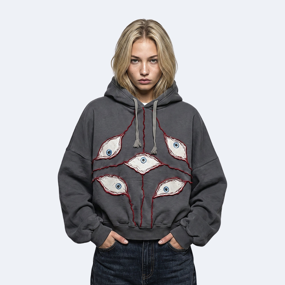 A woman wearing a dark gray oversized hoodie with a graphic of five stylized eyes arranged vertically on the front, paired with dark blue jeans, against a light gray background.