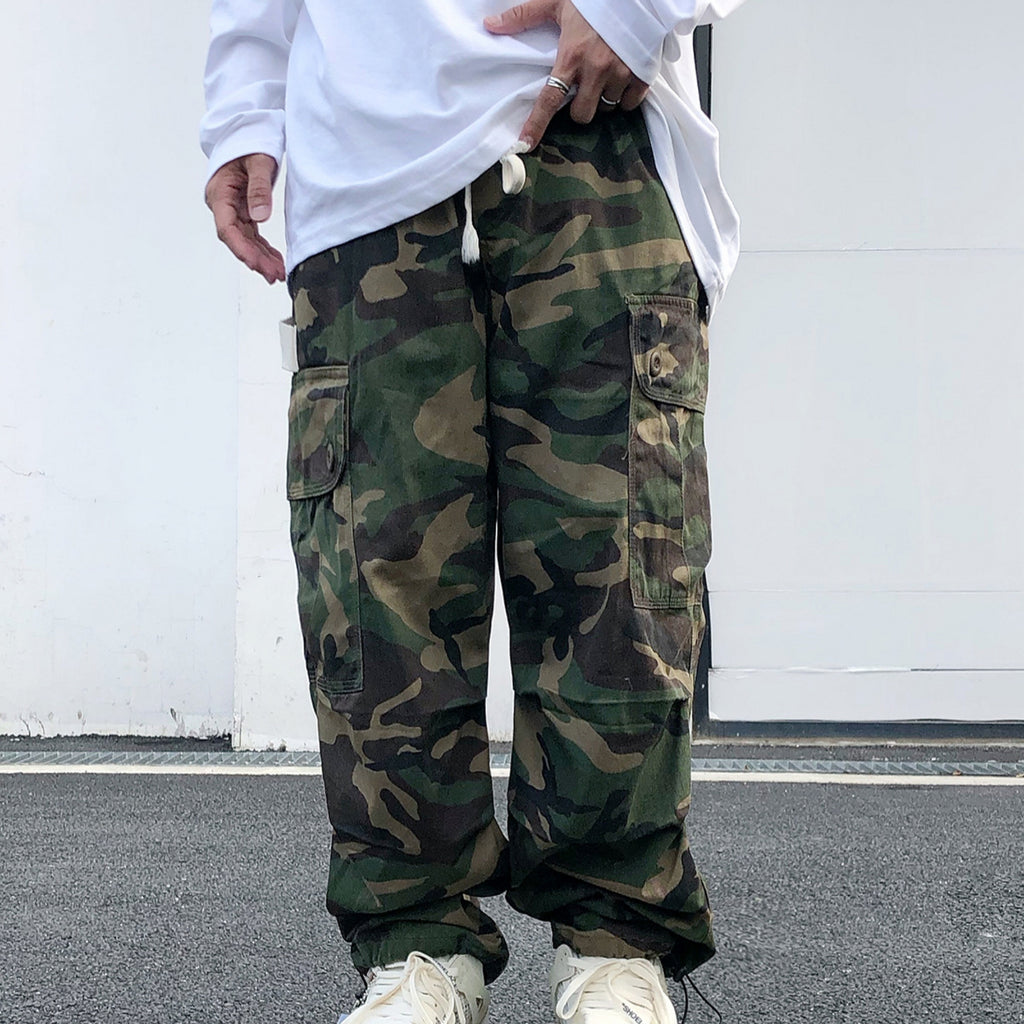 Person wearing camouflage cargo pants with a white shirt and white sneakers on a plain background