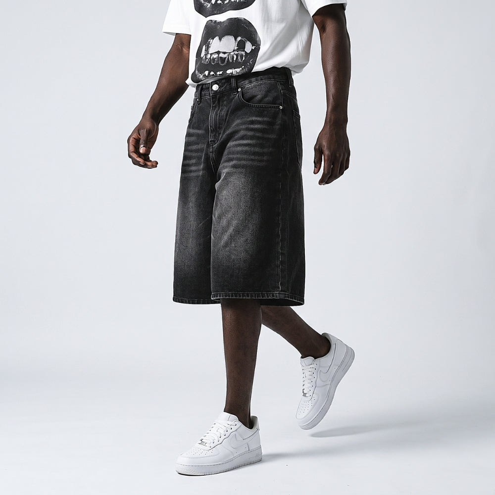 A model wearing black washed denim shorts, a white graphic t-shirt, and white sneakers, shown from the chest down against a light gray studio background.