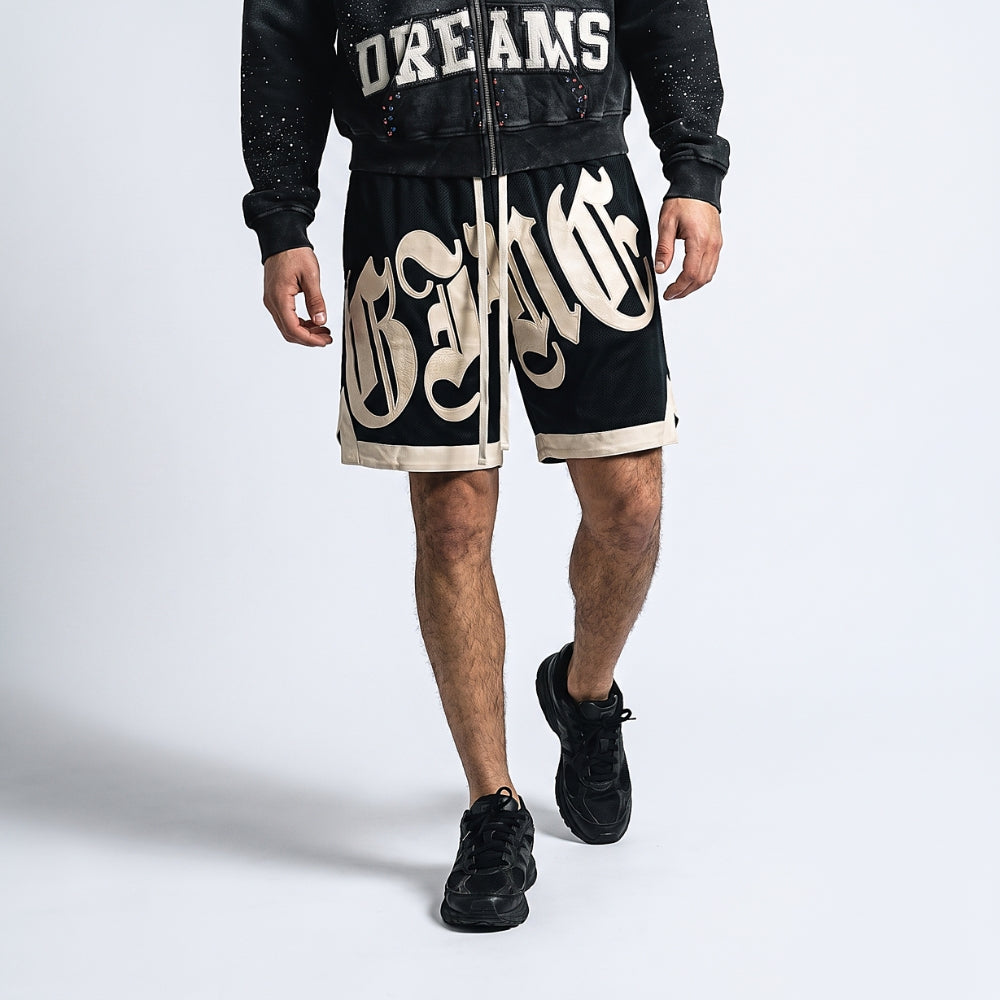 Black. A model wearing black athletic shorts with a large cream gothic-style graphic print, paired with a matching black jacket and black sneakers.
