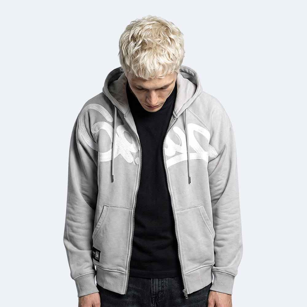 A man with platinum blonde hair wearing a light gray zip-up hoodie with a large white graphic print on the front, layered over a black t-shirt and dark jeans, set against a solid light gray background.