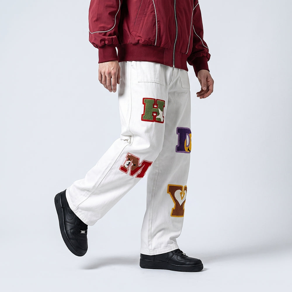 White. A person wearing white varsity-style pants with colorful embroidered letters and patches, paired with a burgundy jacket and black shoes.