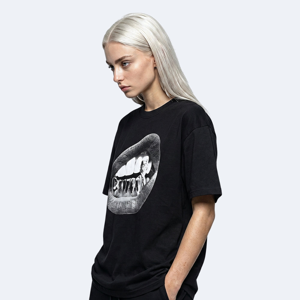 Black. A model with platinum blonde hair wearing a black oversized t-shirt featuring a large black and white graphic of lips and teeth with the word CARNIVAL printed inside.