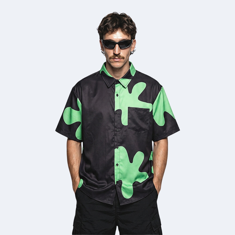 Black & Green. A man wearing a black short-sleeve button-up shirt with a bright green abstract camouflage pattern, styled with black cargo pants and sunglasses.