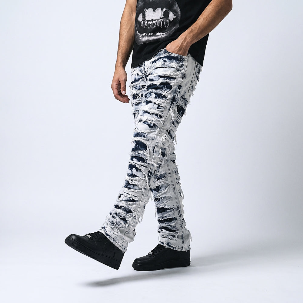 A model wearing distressed, ripped silver and black jeans, a black graphic t-shirt, and black sneakers, captured from the chest down against a light gray studio background.