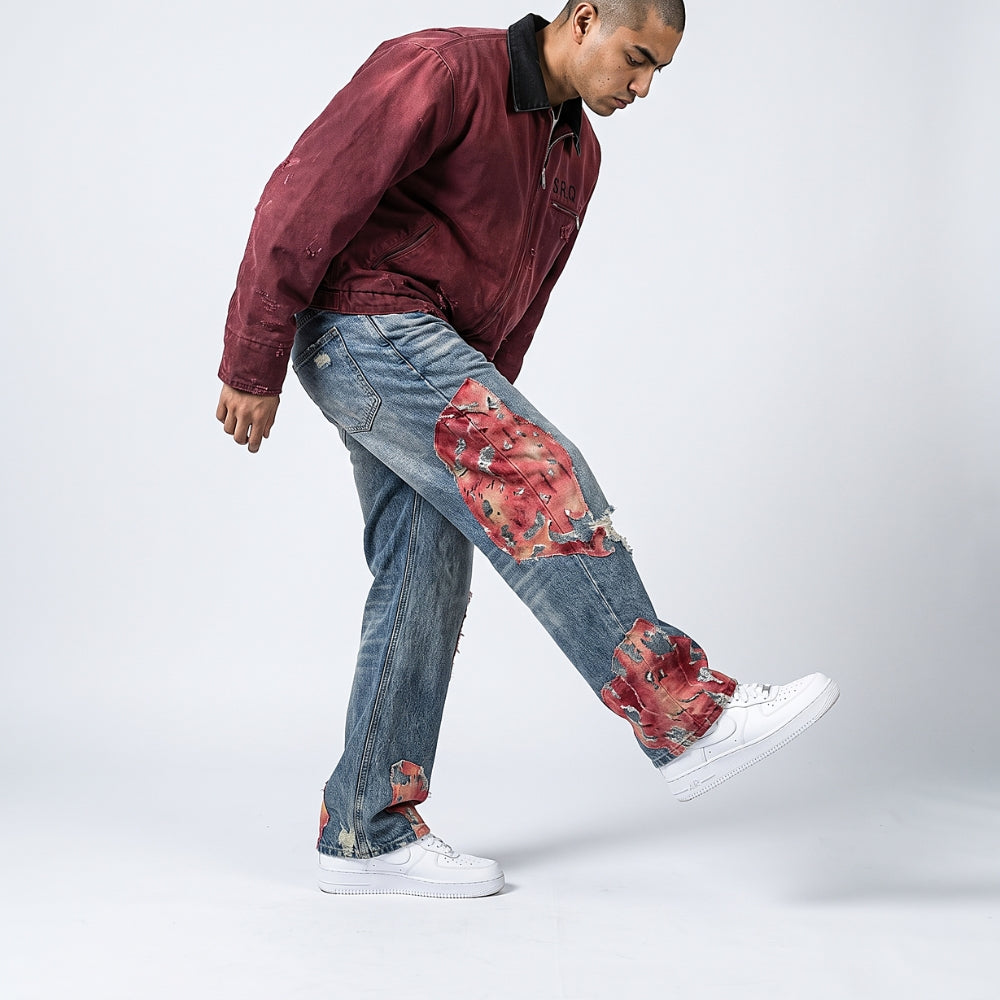 A man wearing a maroon jacket, distressed blue jeans with red floral patchwork details, and white sneakers, captured in a dynamic pose against a light gray studio background.