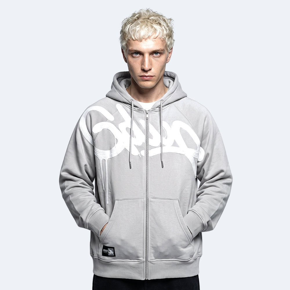 A man with platinum blonde hair wearing a light gray zip-up hoodie with a large white graffiti-style graphic across the chest, posed against a light gray studio background.