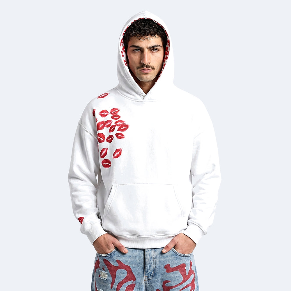 A man wearing a white pullover hoodie with red lip prints on the chest and sleeve, paired with light blue jeans featuring red patterns, set against a plain light gray background.